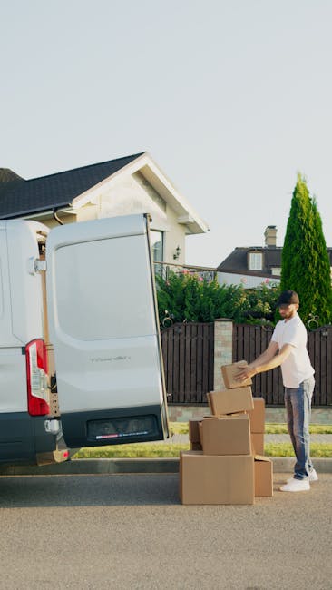 A man wearing a white t-shirt and jeans is standing on a residential street, unloading cardboard boxes from the back of a white van, which is parked at the curb outside a house with a dark roof and white siding. He is placing the boxes on the pavement nearby while the van's rear doors are open, revealing a spacious cargo area ready for home relocation or furniture transport. The background includes a garden with green bushes, a tall conifer tree, and neighboring houses with chimneys and pitched roofs under a clear, daytime sky. This image illustrates the loading process during a professional removals service, highlighting effective packing and handling of belongings for a house move, in alignment with services offered by West Hampstead Man and Van.