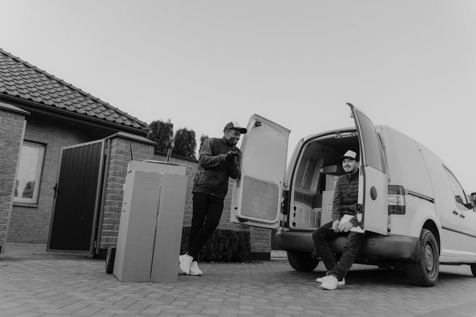 Two movers from West Hampstead Man and Van are engaged in a home relocation process outside a residential property. One individual is standing beside the open rear door of a white delivery van, which is parked on a paved driveway next to a house with a tiled roof and brick walls. The seated mover, wearing a hat and casual clothing, is resting on the edge of the van's open door while smiling and looking towards the standing person. In front of the van, there is a large cardboard box and a black plastic crate, likely containing household items or packed belongings prepared for furniture transport. The scene is set during daylight with clear weather, and inside the property, a window with partially visible curtains can be seen, suggesting the interior space is ready for packing and moving. This image illustrates elements of packing, loading, and vehicle organisation involved in professional removals handled by West Hampstead Man and Van as part of a home relocation service.