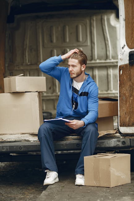 A man wearing a white t-shirt and jeans is standing on a residential street, unloading cardboard boxes from the back of a white van, which is parked at the curb outside a house with a dark roof and white siding. He is placing the boxes on the pavement nearby while the van's rear doors are open, revealing a spacious cargo area ready for home relocation or furniture transport. The background includes a garden with green bushes, a tall conifer tree, and neighboring houses with chimneys and pitched roofs under a clear, daytime sky. This image illustrates the loading process during a professional removals service, highlighting effective packing and handling of belongings for a house move, in alignment with services offered by West Hampstead Man and Van.