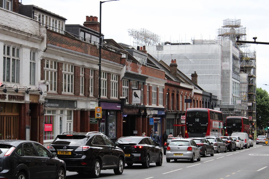 A busy street scene in West Hampstead showing a row of traditional brick terraced houses with white window frames and decorative details. The ground floors of these buildings are occupied by shops, including a café and a fishmonger, with some shop signs visible. Several cars, including black, grey, and white models, are parked along the curb and are in motion on the street. Two red double-decker buses are also present, one approaching and one further down the road, indicating public transport access. The pavement is active with pedestrians walking, some entering or exiting shops. In the background, there is a building under construction, covered in scaffolding and white protective sheeting, suggesting ongoing development work. The street is well-lit with daylight, and the overall scene captures the typical urban environment involved in a home relocation or moving process, as handled by West Hampstead Man and Van, with a focus on transport, loading, and urban logistics.