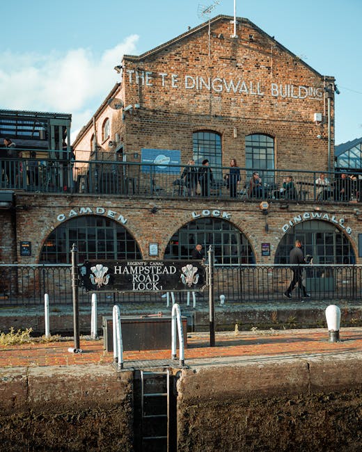 A multi-story brick building known as The Tedingwall Building with large arched windows on the lower level and smaller rectangular windows on the upper level. There is a metal balcony on the top level with a railing, where several people are standing and observing. The building's façade displays signs for Camden Lock and Hampstead Road Lock, indicating its historical significance related to canal lock systems. In front of the building, a black metal fence separates the walkway from a canal or waterway area, with a sign for Hampstead Road Lock and the Regent's Canal. The area is paved with brick and concrete, and there are white bollards along the pavement edge. The scene appears to be during daylight with a partly cloudy sky, and the environment suggests a popular spot for visitors and locals involved in canal and waterfront activities. This setting provides context for an urban outdoor activity that could relate to home relocation logistics, including move planning related to nearby properties or moving equipment. West Hampstead Man and Van may operate in similar urban environments involving house removals or furniture transport near canal or waterway areas.