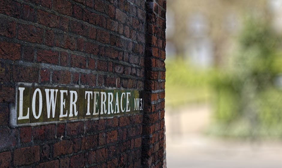 A close-up view of a dark red brick wall with a street sign attached, reading 'LOWER TERRACE NW3' in white capital letters on green and beige background tiles. The sign is positioned on the left side of the image, partially obscured by the corner of the brick wall. In the background, there is an open space with blurred greenery and trees, indicating an outdoor environment. The scene is well-lit with natural daylight, and the image appears to be taken in a residential area, possibly during a home relocation or moving process, as suggested by the surroundings and the context of house removals and furniture transport, with no visible vehicles or moving equipment in the immediate frame.
