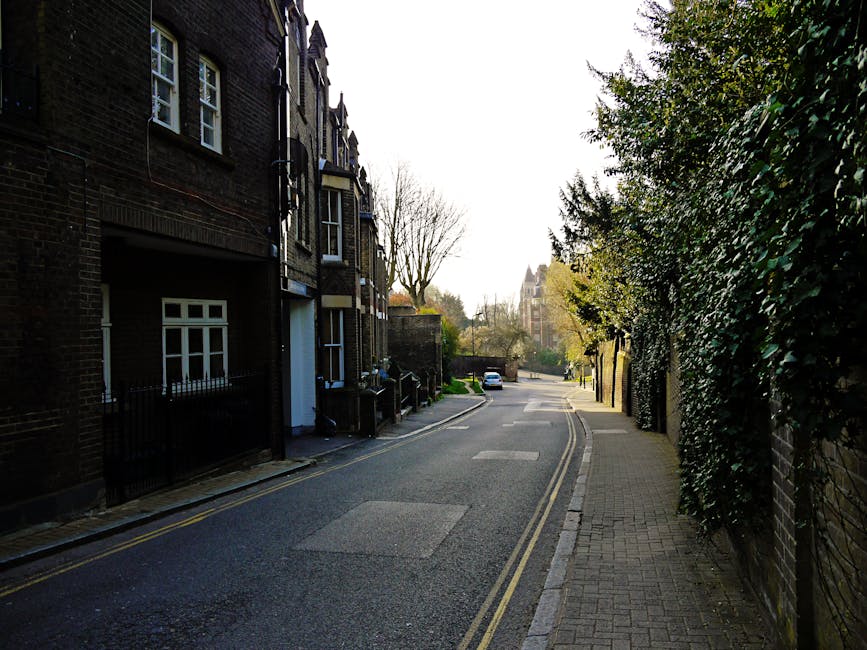 A narrow residential street in West Hampstead with brick terraced houses on the left side and a pavement on the right, lined with green foliage and trees. The street is partly shaded, with a few parked cars visible in the distance. The pavement is made of grey paving stones, and the road has double yellow lines along the curb. This quiet urban environment illustrates typical surroundings for house removals or furniture transport in West Hampstead, with potential access points for moving services like West Hampstead Man and Van, particularly when transporting belongings from flats or houses to vehicles parked nearby.
