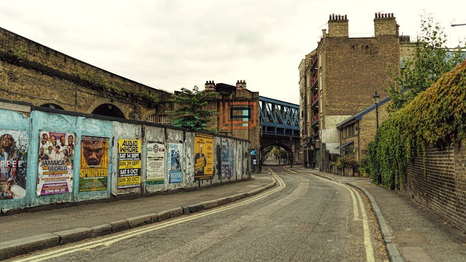 A narrow urban street scene showing a gently curved road with double yellow lines along the curb, leading towards a railway bridge structure painted in blue overhead. On the left side, a tall brick wall is covered with colorful posters advertising local events, with some weathering visible. A pavement runs alongside the wall, and there are no pedestrians present. To the right, a brick building with multiple floors and air conditioning units is bordered by a dense hedge of green foliage extending upward along the wall. The scene is lit by natural diffuse daylight, typical of overcast weather, and the setting suggests a typical area for home relocation or furniture transport, with ample space for loading or unloading near the railway bridge. This environment mirrors a typical location where [COMPANY_NAME] might assist with house and flat removals, especially when navigating through urban streets and access points like those at West Hampstead station to Iverson Road.