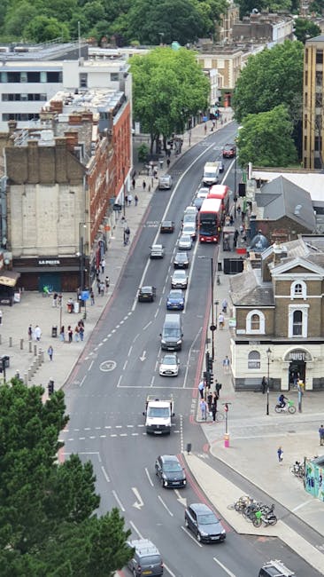 An aerial view of a street in Hampstead Heath with a line of various vehicles, including cars, vans, and a double-decker bus, traveling along the road that curves past a mix of modern and historic buildings. On the left side of the street, pedestrians are walking on the pavement, some near shop entrances, while on the right side, a small church-style building with a grey roof and ornate white trim is visible. Green trees line the sidewalk and the surrounding area, providing a lush backdrop. In the foreground, a fleet of parked bicycles is secured to bike racks, and a few people are seen near the intersections. The scene captures an active urban environment typical of West Hampstead, illustrating a busy city street suitable for home removals, furniture transport, and moving logistics undertaken by West Hampstead Man and Van during packing and loading processes.