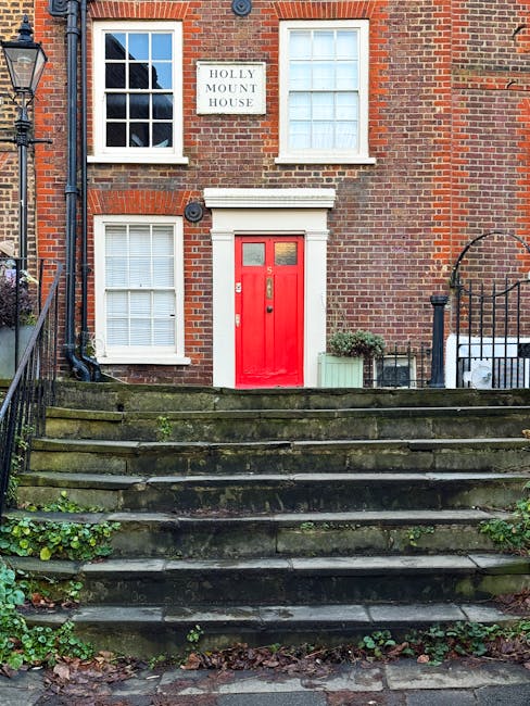 Photo of a weathered brick wall with a black wrought iron grate at the bottom, situated along a street. Above the grate, there is a painted street sign indicating directions to 'Branch Hill' and 'West Heath Road,' with some letters faded or missing. A large tree with moss-covered bark and leafless branches grows behind the wall, extending above it, with parts of nearby residential buildings with brick and tiled roofs visible in the background. The scene is outdoors with natural lighting, and the overall setting suggests a quiet neighbourhood, suitable for a house removal or transport service such as those offered by West Hampstead Man and Van for relocations from Hampstead Heath to Fortune Green.