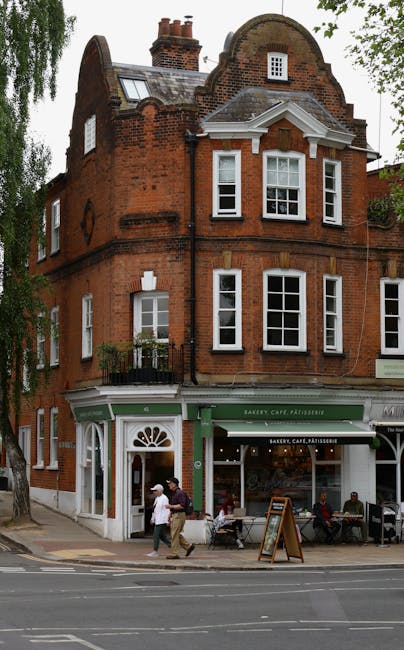 A three-storey red brick building with a curved arch attic and a pitched roof, featuring multiple white-framed sash windows on each floor. The ground floor houses a bakery, café, and pâtisserie with a green storefront, white signage, and large glass display windows. In front of the shop, two men are walking along the sidewalk beside a parked blue van, which is partially visible and used for moving or loading furniture and boxes, with some materials like cardboard boxes and packing supplies nearby. The surrounding environment includes a tree on the left side, and the street has marked lanes and tramlines, indicating a busy urban area suitable for house and office relocations. The scene is set during daylight with overcast weather.